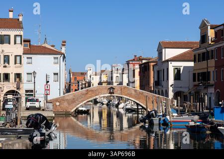 Chioggia, Italia - 3 marzo 2025: Ponte in mattoni sul Canal Vena con barche ormeggiate ed edifici colorati nel centro storico Foto Stock