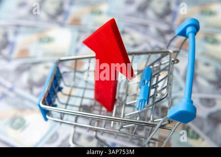 Un carrello della spesa e una freccia rossa rivolta verso l'alto su un letto di banconote sfocate da cento dollari. Aumento dei costi, inflazione. Foto Stock