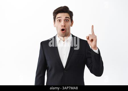 Uomo d'affari sorpreso che punta il dito in su e che gastping stupito, dica WOW, stare eccitato alla macchina fotografica, in piedi in vestito nero contro sfondo bianco dello studio. Foto Stock
