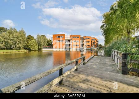 Un elegante e moderno complesso di appartamenti si trova accanto a un tranquillo fiume, circondato da una vegetazione lussureggiante sotto un cielo blu limpido, ideale per vivere sereno in riva all'acqua. Foto Stock
