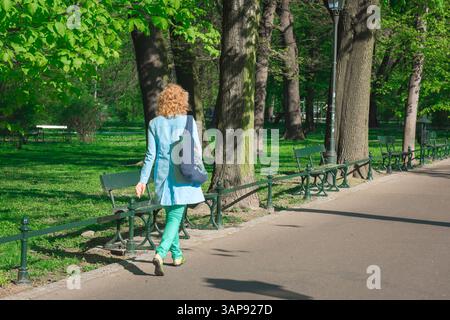 Donna che cammina da sola, veduta posteriore di una giovane donna che indossa un impermeabile blu che cammina attraverso Planty Park nel centro di Cracovia, in Polonia. Foto Stock