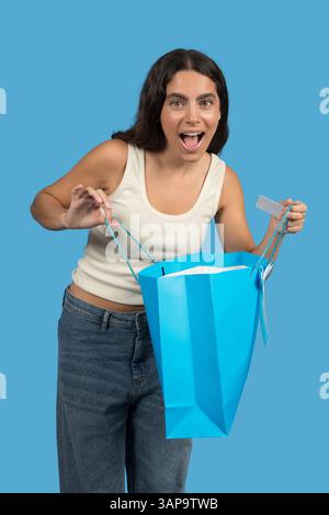 Una giovane donna sembra sorpresa mentre apre una borsa della spesa e mostra una carta fedeltà Foto Stock
