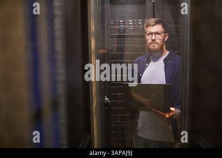 Ritratto del tecnico che si trova nella sala server con un laptop e che gestisce le apparecchiature server. Il tecnico sta guardando la telecamera Foto Stock