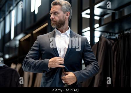 Uomo maturo in un abito in una boutique di abiti. Un uomo d'affari sceglie con fiducia un abito elegante in negozio. Un cliente alla moda esamina la giacca mentre fa shopping Foto Stock