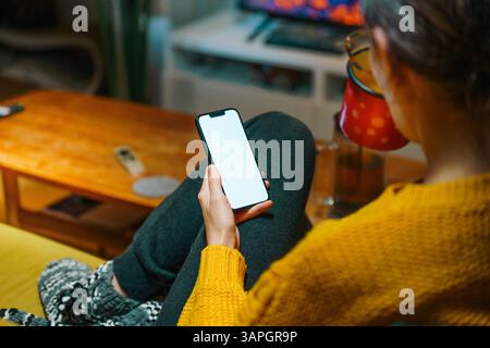Calda atmosfera di casa con donna che usa lo smartphone mentre si rilassa accanto al caminetto, comfort invernale Foto Stock