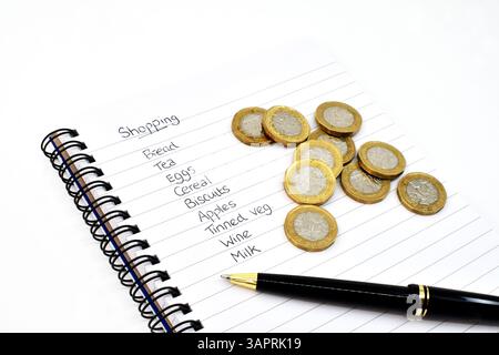 Vista ravvicinata di una lista della spesa scritta su una pagina di un notebool con carta foderata, con monete inglesi da una sterlina e una penna. Niente persone. Foto Stock