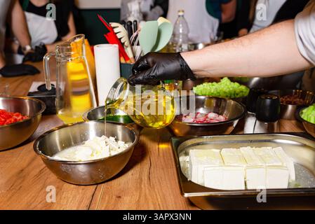 Uno chef versa olio d'oliva dorato sul formaggio feta sbriciolato in una ciotola di metallo, circondata da ingredienti freschi per insalata. Foto Stock