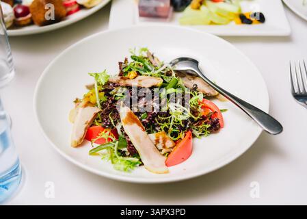 Insalata fresca di verdure a foglia verde, pomodori e fette di pollo alla griglia servita in un recipiente bianco con un cucchiaio su una tovaglia bianca. Foto Stock
