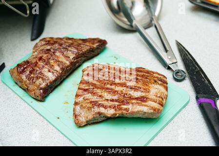 Due bistecche di manzo grigliate con segni di cucitura appoggiate su un tagliere verde, pronte per affettare o servire con i contorni. Foto Stock