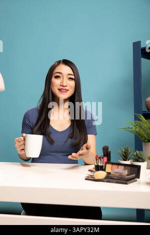 Vlogger di bellezza asiatico, sorridente e parla con i suoi spettatori durante una trasmissione in diretta in uno studio con sfondo blu. Si siede alla sua scrivania, tenendo una tazza di caffè e facendo un contatto visivo con la telecamera. Foto Stock