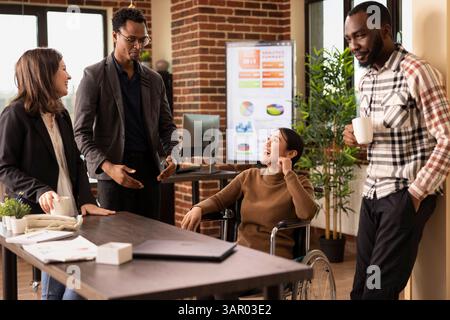 Professionisti multirazziali in una startup creativa che si prendono un momento per rilassarsi, chiacchierare e ridere insieme durante una pausa. Uomo afroamericano con gli occhiali, che condivide una storia con la sua squadra. Foto Stock