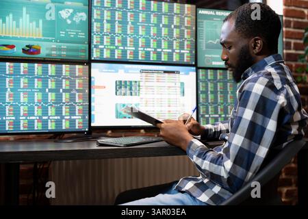 Il manager afroamericano si siede e scrive sugli appunti, registrando gli aggiornamenti dei progressi per gli hedge fund dell'azienda. Professionista maschile che prende appunti, lavora all'analisi del rischio della negoziazione di azioni in ufficio. Foto Stock