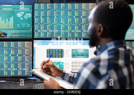Primo piano di un dipendente nero che analizza le tendenze di acquisto e vendita su più monitor e scrive aggiornamenti sugli appunti. L'agente di borsa si è concentrato su una sessione di trading presso la sua postazione di lavoro high-tech. Foto Stock