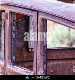 Primo piano di una portiera e dei finestrini di un'auto d'epoca arrugginiti con vetri mancanti, che mostrano dettagli in metallo e legno intemprati in una luce soffusa. Foto Stock