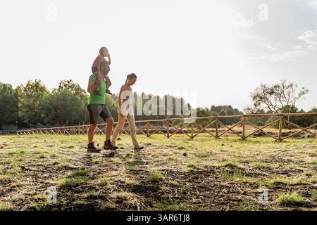 Papà porta sua figlia sulle spalle mentre cammina con la mamma su un soleggiato sentiero rurale. La famiglia si diverte nella natura. Foto Stock