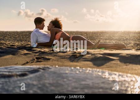 Coppie interrazziali che amano godersi un romantico tramonto sulla spiaggia. Donna che giace sull'uomo, sorridente e rilassata. Vacanze estive e momenti intimi vicino al Foto Stock