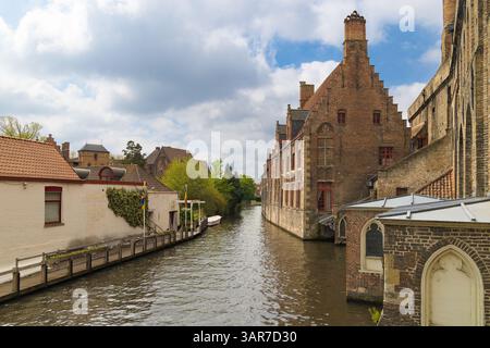 Edifici storici in mattoni, tra cui Sint-Janshospitaal, costeggiano un canale panoramico a Bruges, Belgio, sotto un cielo parzialmente nuvoloso. L'acqua riflette gli edifici. Foto Stock