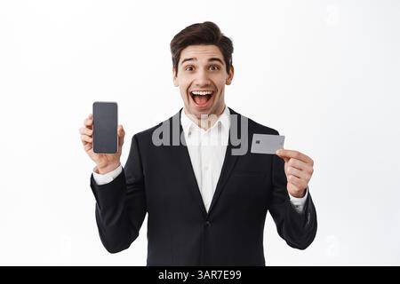 Uomo d'affari sorpreso, uomo d'affari in vestito mostra schermo del telefono vuoto e carta di credito, sorridente stupito, nuova app di banca, in piedi su sfondo bianco. Foto Stock