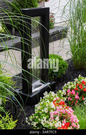 Cornice metallica rettangolare nera illuminata ornamento di fontane delimitato da arbusti Thuja occidentalis e fiori di Begonia nel giardino frontale in estate. Foto Stock