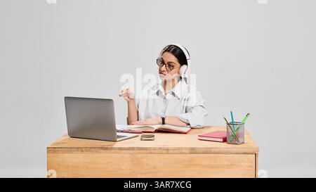 Insegnante donna concentrata in camicia bianca e cuffie che spiega l'argomento durante la lezione online con gesti espressivi. Foto Stock