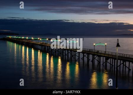 Un lungo molo di legno si estende su acque tranquille, adornato da luci accese al tramonto. I riflessi risplendono sulla superficie, migliorando la serenità Foto Stock
