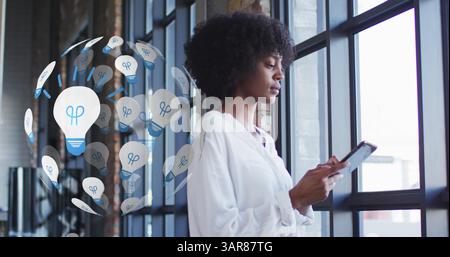 Immagine di un globo di icone di lampadine sopra una donna afroamericana che usa un tablet Foto Stock