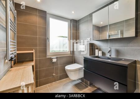 Un bagno elegante e moderno caratterizzato da un'estetica minimalista con piastrelle scure, un lavandino contemporaneo e luce naturale da una grande finestra. Foto Stock