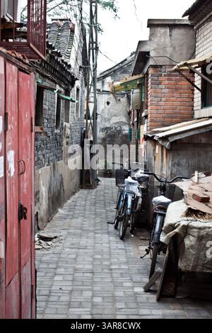 Pechino, Cina, 14.07.2010. Un vicolo stretto con una porta rossa e alcune biciclette parcheggiate sul marciapiede. Le moto sono incatenate a un muro e a una panchina i. Foto Stock
