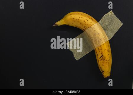 Una banana matura è fissata con un pezzo di nastro trasparente su un solido sfondo nero, creando un contrasto sorprendente e una presentazione insolita Foto Stock