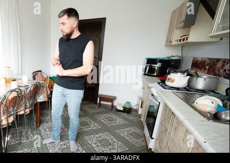 Un uomo con un piano senza maniche si trova in una cucina luminosa, impegnata nella preparazione dei pasti, circondato da un'atmosfera accogliente e cucina quotidiana Foto Stock
