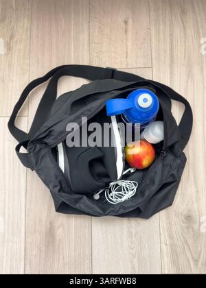 Una borsa nera contenente scarpe da ginnastica, cuffie, una mela, un deodorante e una bottiglia d'acqua. Set di attrezzature sportive sul pavimento. Fitness, palestra, allenamento. Foto Stock