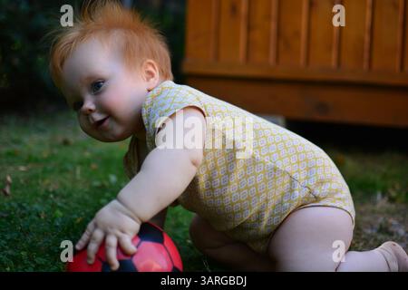 La bambina dai capelli rossi in tuta gialla gioca in un cortile posteriore con palla da calcio rossa e nera. Bimbo sorridente e strisciante sull'erba fuori da Foto Stock