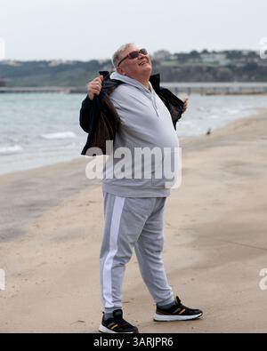 Uomo anziano che abbraccia la brezza marina con gioia su una spiaggia tranquilla, indossa abbigliamento sportivo casual, occhiali da sole e un sorriso leggero Foto Stock