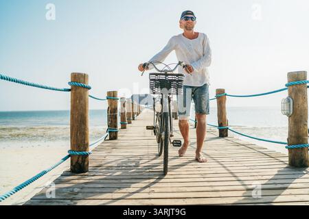 Ritratto dell'uomo vestito con leggeri abiti estivi, occhiali da sole con vecchia bicicletta sul molo di legno sulla spiaggia sabbiosa di Zanzibar. Vacanza disinteressata in t Foto Stock