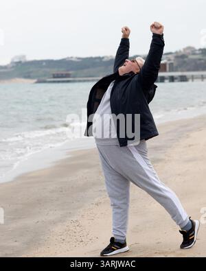 Un uomo anziano gioioso che celebra la vita su una spiaggia ghiacciata, indossa una giacca scura, pantaloni tuta grigi e sneakers nere, che si allunga verso l'alto con le braccia Foto Stock