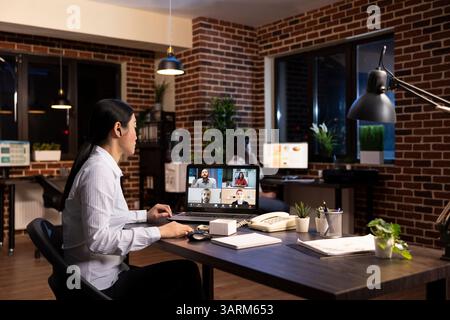 Una manager asiatica si impegna in una riunione virtuale con il suo team, condividendo idee di strategia aziendale durante una videoconferenza. Il laptop mostra che si connette con i dipendenti remoti durante il turno di notte. Foto Stock