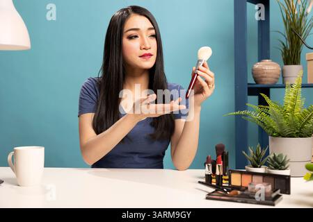 Vlogger di bellezza che spiega i suggerimenti per il trucco tenendo in mano una spazzola. Donna asiatica seduta alla sua scrivania, gestando verso lo strumento cosmetico, facendo un video tutorial e connettendosi con i suoi seguaci online. Foto Stock