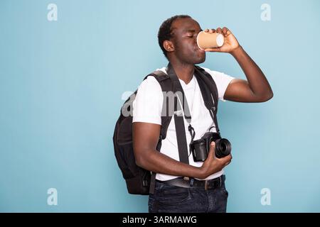 Fotografo nero in abbigliamento casual con zaino e una bevanda calda. Un afroamericano con una macchina fotografica intorno al collo sorseggia un caffè mentre si trova su uno sfondo isolato. Foto Stock