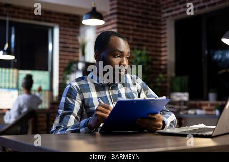 Il manager afroamericano si siede alla sua scrivania, raccogliendo gli aggiornamenti sui progressi su un appunti per l'hedge fund dell'azienda. Consulente finanziario prende appunti mentre analizza i rischi di negoziazione delle azioni in ufficio. Foto Stock