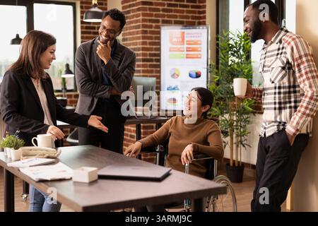 Donna d'affari in sedia a rotelle ha partecipato a una discussione amichevole con i suoi colleghi, evidenziando un luogo di lavoro accessibile e diversificato. Donna caucasica che racconta una storia divertente ai suoi colleghi durante la pausa. Foto Stock