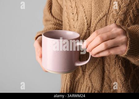 Donna in un elegante pullover lavorato a maglia con tazza in ceramica su sfondo grigio, primo piano Foto Stock