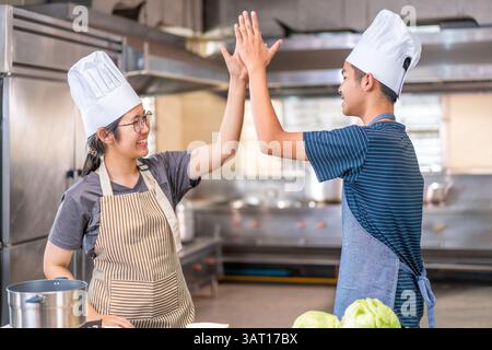 Due giovani chef festeggiano con un alto cinque in una cucina commerciale, indossando cappelli e grembiuli da chef, mostrando lavoro di squadra e gioia nella cucina. Foto Stock