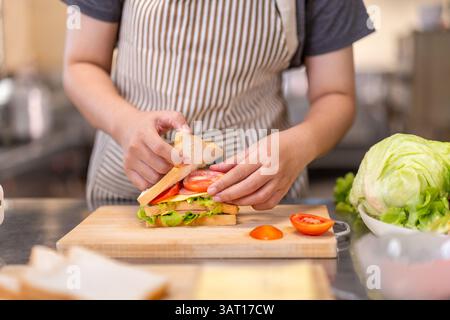 Un primo piano delle mani di uno chef che assembla un panino fresco con strati di lattuga, uova e formaggio su un tagliere di legno. I pomodori a fette lo sono Foto Stock
