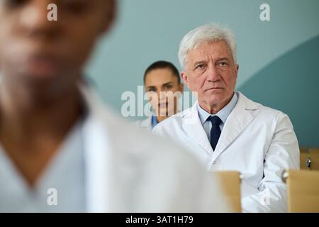 Medico senior maschile e giovane medico donna in camici da laboratorio bianchi ascoltando attentamente durante una conversazione critica in ambiente ospedaliero, sfondo sli Foto Stock