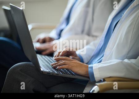 I medici che lavorano su computer portatili durante la consultazione in ambiente ospedaliero, indossano camici bianchi e si concentrano sulle cartelle dei pazienti, sulla digitazione delle informazioni e sul collabo Foto Stock