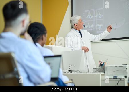 Scienziato senior che tiene lezioni in classe moderna utilizzando schermo e tecnologia, ascoltando e prendendo appunti con attenzione da un pubblico eterogeneo Foto Stock