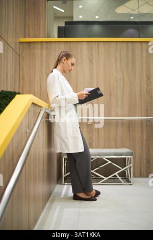 Medico in piedi nel corridoio, rivedendo documenti importanti sugli appunti mentre si appoggia contro un muro di legno, concentrandosi sul suo lavoro con de Foto Stock