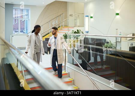 Due professionisti medici conversano mentre camminano giù per le scale in ambiente ospedaliero, indossando camici da laboratorio, concentrandosi sulle opzioni di cura e trattamento del paziente Foto Stock