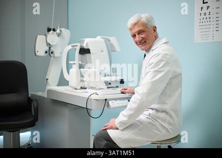Medico anziano sorridente calorosamente mentre si siede in una clinica oculistica moderna, circondato da attrezzature mediche avanzate Foto Stock