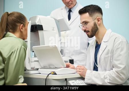 Donna sottoposta a un esame oculistico con un optometrista che utilizza apparecchiature mediche in ambiente clinico. Medico di alto livello che osserva la procedura in background Foto Stock